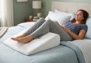 Person lying on their back with legs resting on a white foam wedge leg elevation pillow on a bed